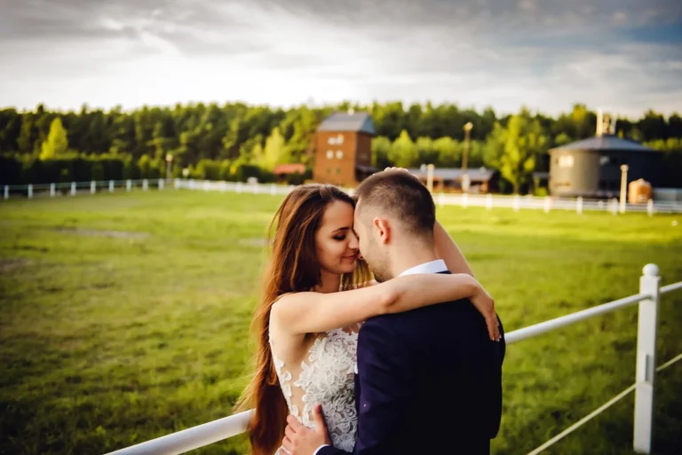 Jak wybrać fotografa ślubnego i porównać pakiety? Para młoda w objęciach przy białym ogrodzeniu